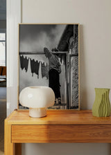 Quadro fotográfico de uma criança de costas sobre uma estrutura metálica, em tons de preto, cinzento e branco, com fachadas urbanas, roupa estendida e estilo documental nostálgico. Disponível em kudeko.com