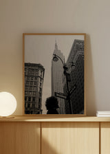 Quadro emoldurado com cena urbana de Nova Iorque a preto e branco, Empire State Building, candeeiros pretos, edifícios históricos, placa de Broadway e silhueta humana em primeiro plano, num estilo citadino contemporâneo. Disponível em kudeko.com