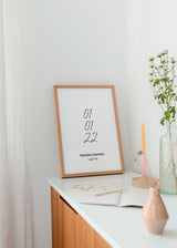 Tableau encadré personnalisé avec date en chiffres noirs, prénoms et lieu sur fond blanc, composition épurée au style minimaliste pour une décoration murale sobre. Disponible sur kudeko.com