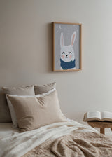 Gerahmtes Wandbild mit weißem Hasen, rosigen Wangen, blauem Schal, grauem Schneehintergrund, Mond, Sternen und kindlichem Stil. Erhältlich auf kudeko.com