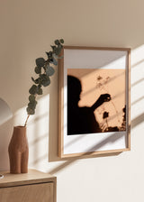 Quadro com silhueta feminina sombreada a segurar flores silvestres, fundo bege quente, contraste preto e estilo fotográfico poético. Disponível em kudeko.com