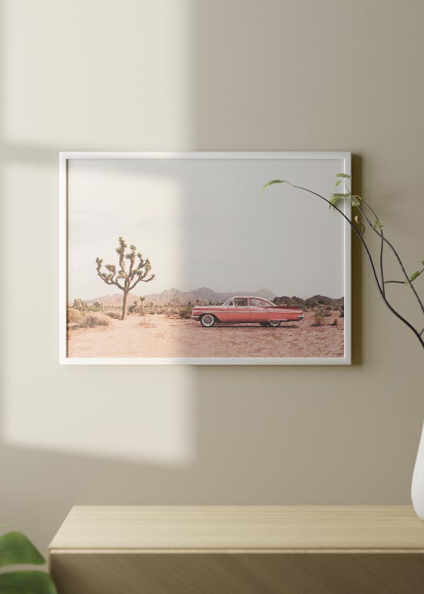 Horizontales Wandbild mit rosafarbenem Oldtimer in Wüstenlandschaft, beigefarbenem Sand, trockenen Sträuchern, Joshua Tree, sanften Bergen und fotografischem Vintage-Stil in pudrigen Tönen. Erhältlich auf kudeko.com