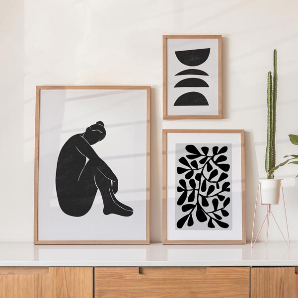 Set aus drei Wandbildern mit sitzender abstrakter Frauenfigur, schwarzen halbkreisförmigen geometrischen Formen, geschwungenen Blättern auf weißem Hintergrund im minimalistischen Stil. Erhältlich auf kudeko.com