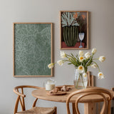 Tableau avec vase cannelé vert olive, branches vert clair, petites fleurs roses, deux fleurs orange, verre blanc, fond brun et style floral contemporain. Disponible sur kudeko.com