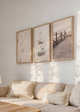 Conjunto de três quadros com conchas marinhas, passadiço de madeira junto ao mar, areia clara, rosa velho, branco, bege suave e estilo fotográfico costeiro. Disponível em kudeko.com