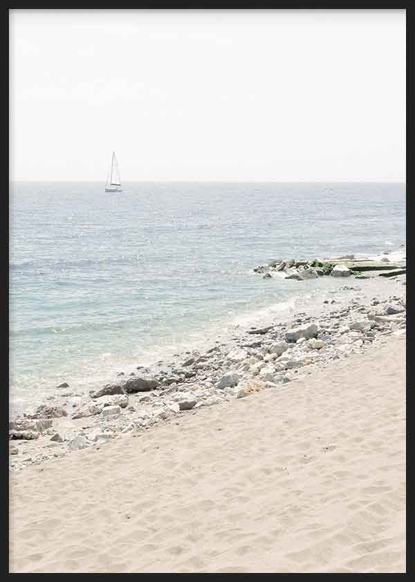 Cuadro de playa luminosa con arena beige, orilla de piedras claras, mar azul suave y velero lejano, de estilo fotográfico. Disponible en kudeko.com