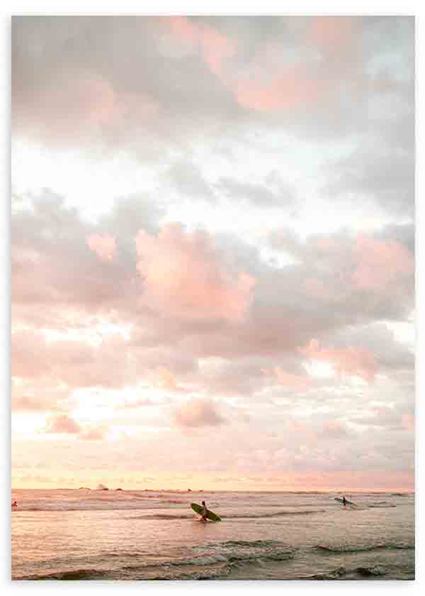 Quadro emoldurado com dois surfistas no mar ao pôr do sol, céu com nuvens rosa, cinzento suave e creme, horizonte delicado e estilo fotográfico sereno. Disponível em kudeko.com
