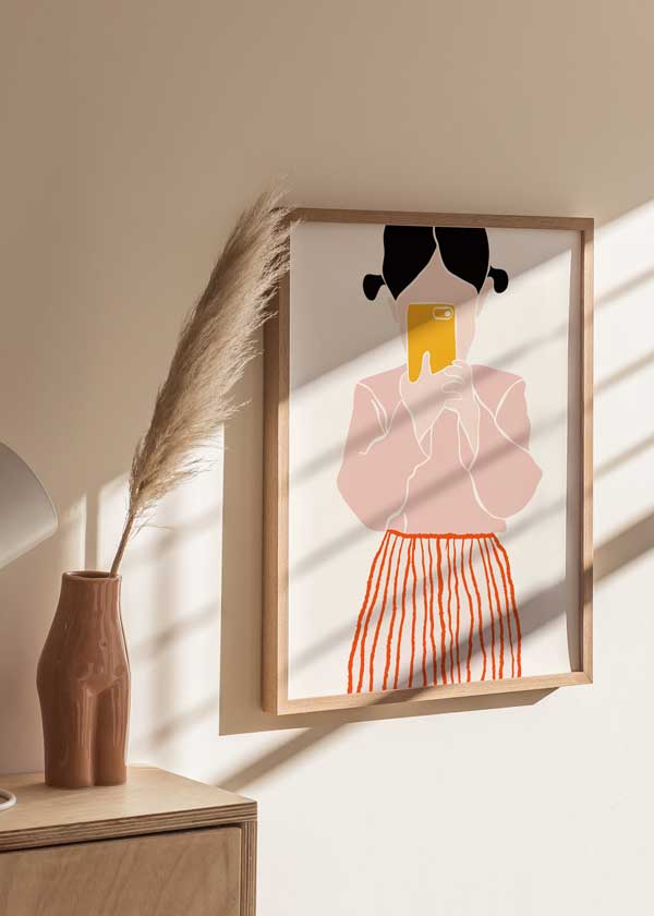 Tableau encadré d’une fillette avec couettes, téléphone jaune devant le visage, blouse rose, jupe à rayures orange, fond clair et style d’illustration enfantine. Disponible sur kudeko.com