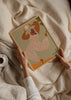 Wandbild mit stilisierter weiblicher Figur, weißem Overall mit korallfarbenen Streifen, heller Sonne, beigem Hintergrund, grünen Details und zeitgemäßem Illustrationsstil mit organischen Formen. Erhältlich auf kudeko.com