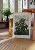 Quadro botânico com begónia, folhas grandes verde escuro, caules avermelhados, flores em rosa claro e branco sobre fundo verde acinzentado, estilo vintage. Disponível em kudeko.com