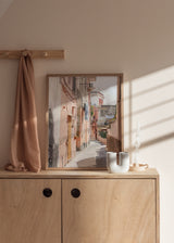 Quadro emoldurado de uma rua estreita mediterrânica com fachadas em rosa, bege e terracota, chão empedrado, varandas, roupa estendida, luz suave e estilo fotográfico. Disponível em kudeko.com