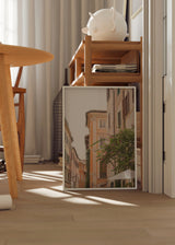 Gerahmtes Wandbild einer römischen Straße mit Fassaden in Creme, Pfirsich und Sand, dunklen Fensterläden, Balkonen, grüner Vegetation, fuchsiafarbenen Blüten und mediterranem Stadtstil. Erhältlich auf kudeko.com