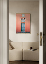 Fotografisches Wandbild mit verwitterter blauer Tür auf korallfarbener Fassade, Fenster mit doppelten Bögen, türkisen und weißen Details, symmetrischer Komposition und mediterranem Architekturstil. Erhältlich auf kudeko.com
