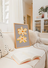Tableau encadré avec deux grandes fleurs crème, pêche, orange et jaune sur fond gris taupe, dans un style illustration moderne et épuré. Disponible sur kudeko.com