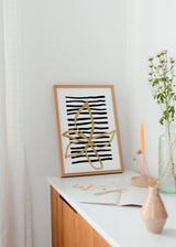Gerahmtes Wandbild mit großer goldener Linienblüte, unregelmäßigen schwarzen Streifen, weißem Hintergrund und minimalistischem zeitgenössischem Stil. Erhältlich auf kudeko.com