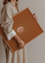 Gerahmtes Wandbild mit abstrakter Sonne, rosa Mittelpunkt, feinen cremefarbenen Strahlen, terrakottafarbenem Hintergrund und minimalistischer, ausgewogener Gestaltung. Erhältlich auf kudeko.com