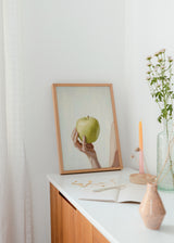 Quadro minimalista de uma mão segurando uma maçã verde. Disponível em kudeko.com.