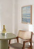 Quadro emoldurado de praia com ondas suaves, céu nublado, figura solitária junto à beira-mar, em azul-claro, branco e cinzento, de estilo vintage. Disponível em kudeko.com