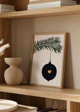 Quadro com bola de Natal preta suspensa, pequeno coração dourado, ramo de pinheiro verde, fundo claro e estilo minimalista para decoração de parede festiva. Disponível em kudeko.com