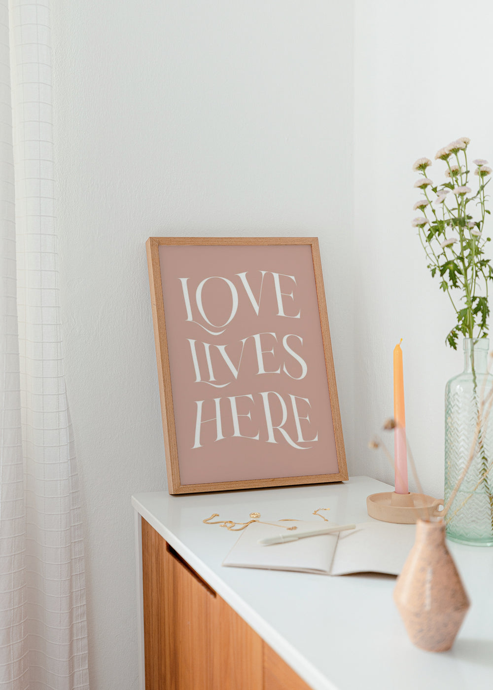 Tableau encadré avec la phrase LOVE LIVES HERE, grandes lettres blanches sur fond rose poudré, composition typographique élégante dans un style décoratif à message. Disponible sur kudeko.com