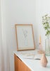 Gerahmtes Wandbild mit weißer Vase in organischer Form, feinen Blumenlinien, zarten Stielen, beigem und cremeweißem Hintergrund, botanische Komposition im minimalistischen Stil. Erhältlich auf kudeko.com