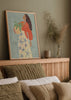Tableau encadré avec femme de profil tenant une plante en pot, fond bleu gris doux, robe crème à fleurs bleues, blouse terracotta, style illustration décorative. Disponible sur kudeko.com