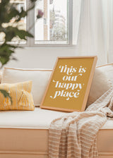 Gerahmtes Wandbild mit dem Spruch This is our happy place in großer weißer Typografie auf senfgelbem Hintergrund, im modernen und reduzierten Schriftstil. Erhältlich auf kudeko.com