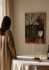 Tableau avec vase cannelé vert olive, branches vert clair, petites fleurs roses, deux fleurs orange, verre blanc, fond brun et style floral contemporain. Disponible sur kudeko.com