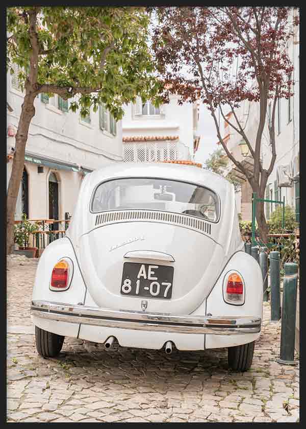 Quadro com carro clássico branco numa rua empedrada, com árvores verdes, fachadas claras e ambiente mediterrânico, em estilo fotográfico vintage. Disponível em kudeko.com