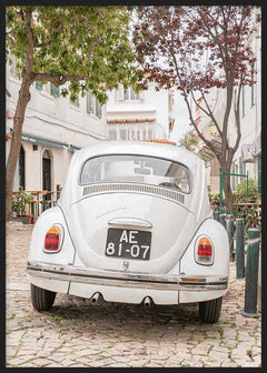 Quadro com carro clássico branco numa rua empedrada, com árvores verdes, fachadas claras e ambiente mediterrânico, em estilo fotográfico vintage. Disponível em kudeko.com