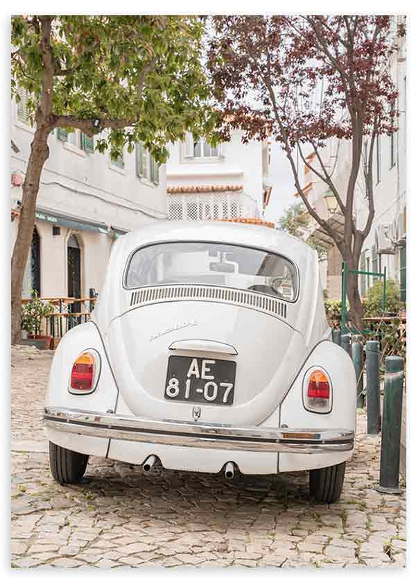 Quadro com carro clássico branco numa rua empedrada, com árvores verdes, fachadas claras e ambiente mediterrânico, em estilo fotográfico vintage. Disponível em kudeko.com