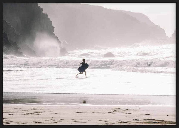 Cuadro horizontal de surfista caminando hacia el mar entre olas blancas, arena húmeda, acantilados grises y luz suave, de estilo fotográfico. Disponible en kudeko.com