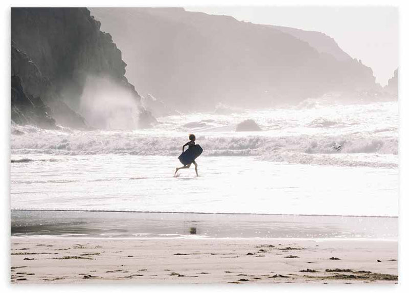 Cuadro horizontal de surfista caminando hacia el mar entre olas blancas, arena húmeda, acantilados grises y luz suave, de estilo fotográfico. Disponible en kudeko.com