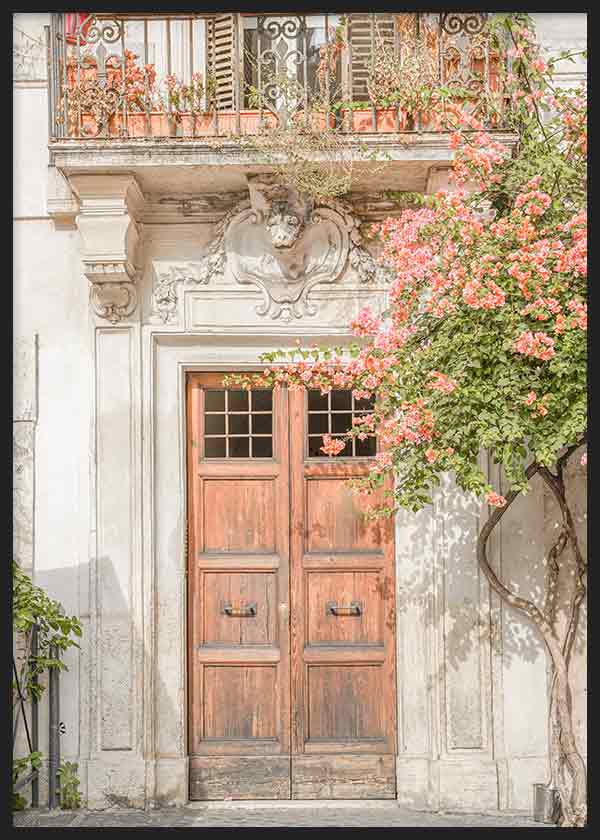Cuadro de entrada clásica con puerta de madera, balcón ornamental, flores rosadas y vegetación verde sobre fachada en tonos crema, de estilo ciudades. Disponible en kudeko.com