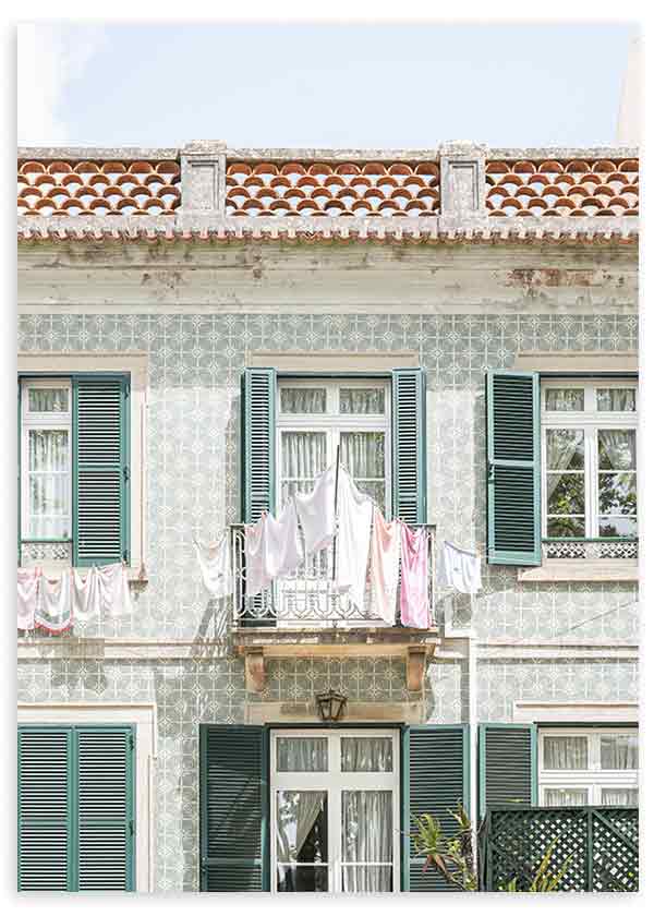 Quadro de fachada portuguesa com azulejos claros, varanda central, roupa estendida, janelas com portadas verdes e telhado terracota, num estilo fotográfico mediterrânico. Disponível em kudeko.com