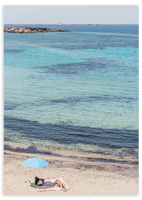 Cuadro de playa mediterránea con mar turquesa, arena clara, sombrilla azul y dos personas descansando, de estilo fotográfico. Disponible en kudeko.com