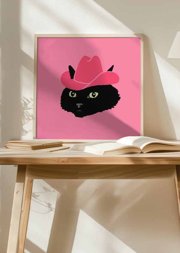 Cuadro cuadrado con gato negro de ojos amarillo verdoso y sombrero vaquero rosa sobre fondo rosa, en estilo ilustración. Disponible en kudeko.com