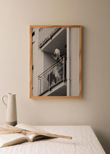 Cuadro de mujer en balcón de edificio urbano, en tonos blanco y negro, con barandillas metálicas, escena cotidiana y estilo fotográfico. Disponible en kudeko.com