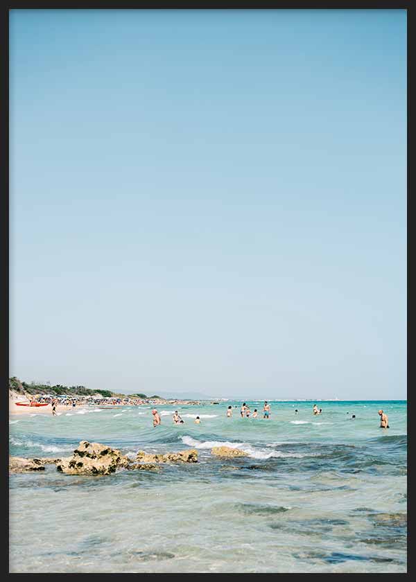 Cuadro de playa mediterránea con bañistas, rocas junto a la orilla, agua turquesa y cielo azul amplio, de estilo fotográfico. Disponible en kudeko.com