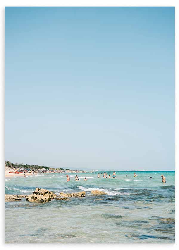 Cuadro de playa mediterránea con bañistas, rocas junto a la orilla, agua turquesa y cielo azul amplio, de estilo fotográfico. Disponible en kudeko.com
