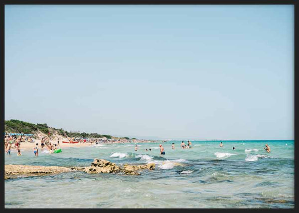 Cuadro horizontal de playa mediterránea con agua turquesa, arena clara, rocas suaves, bañistas dispersos y cielo azul despejado, de estilo fotográfico. Disponible en kudeko.com
