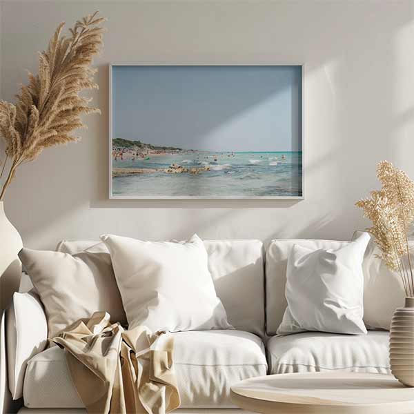 Quadro horizontal de praia mediterrânica com água turquesa, areia clara, pequenas rochas, banhistas dispersos, céu azul suave e estilo fotográfico natural. Disponível em kudeko.com