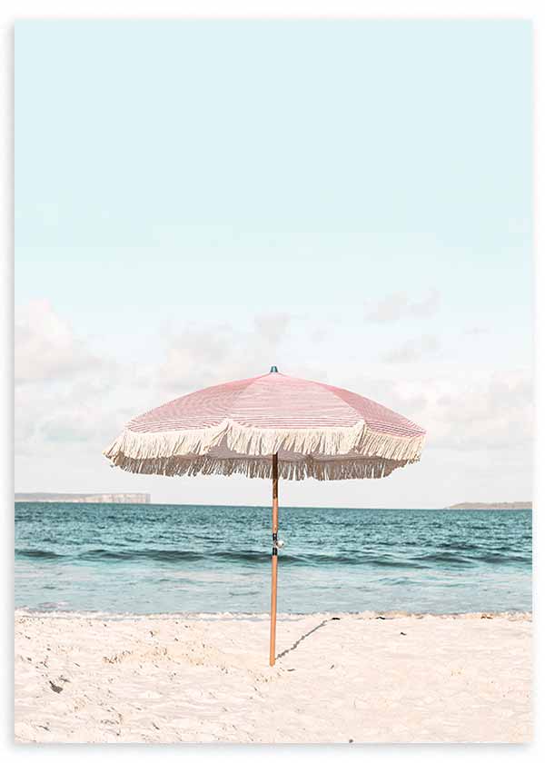 Gerahmtes Wandbild mit rosa Sonnenschirm mit Fransen auf hellem Sandstrand, ruhigem blauem Meer, pastellfarbenem Himmel und hellem Fotostil. Erhältlich auf kudeko.com