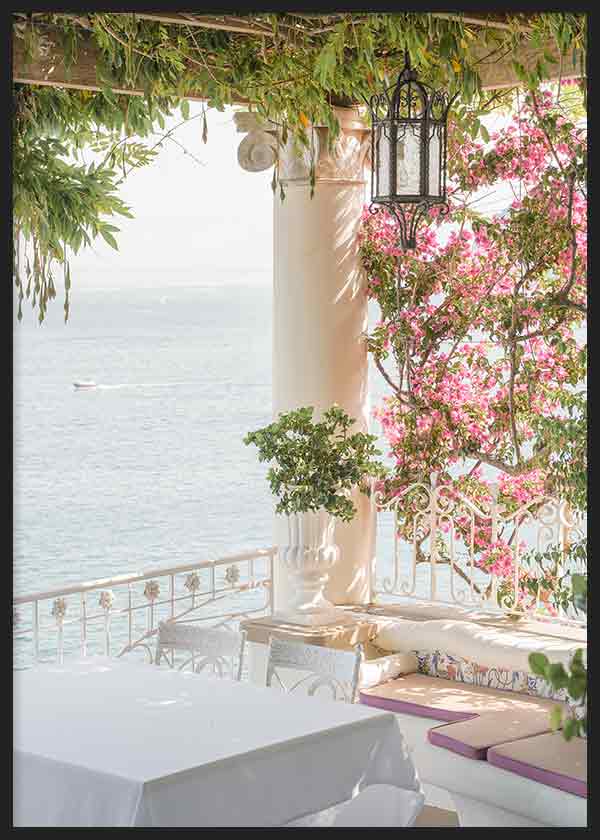 Quadro de terraço mediterrânico junto ao mar com buganvílias cor-de-rosa, colunas brancas, candeeiro preto, vegetação verde e luz suave, em estilo fotográfico luminoso. Disponível em kudeko.com