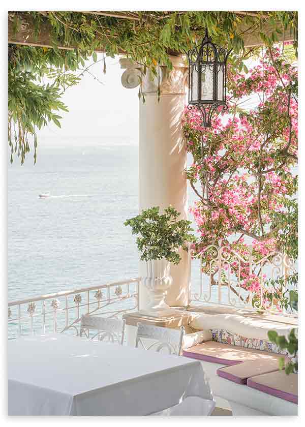 Quadro de terraço mediterrânico junto ao mar com buganvílias cor-de-rosa, colunas brancas, candeeiro preto, vegetação verde e luz suave, em estilo fotográfico luminoso. Disponível em kudeko.com