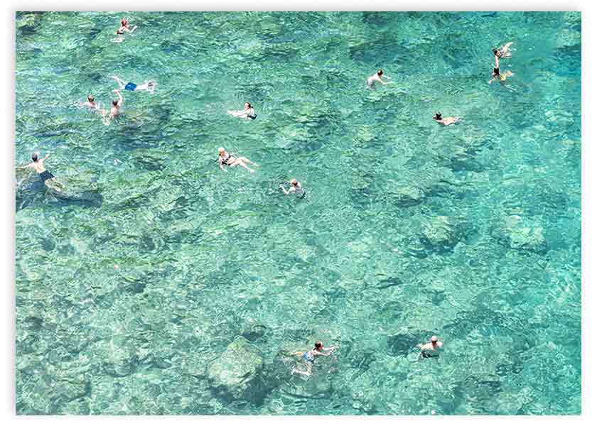 Cuadro horizontal de personas nadando en mar cristalino turquesa y aguamarina, con fondo rocoso visible y estilo fotográfico de aire veraniego. Disponible en kudeko.com