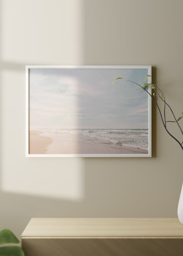 Cuadro horizontal de playa tranquila con orilla de arena clara, olas suaves, horizonte marino y cielo amplio en tonos azul grisáceo, blanco y beige rosado, de estilo fotográfico. Disponible en kudeko.com