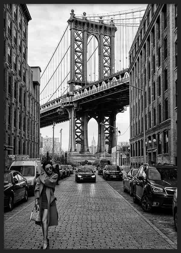 Quadro de rua urbana a preto e branco com grande ponte suspensa, edifícios de tijolo, carros estacionados e figura feminina em primeiro plano, em estilo fotográfico cinematográfico. Disponível em kudeko.com