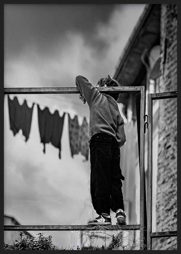 Tableau photographique d’un enfant vu de dos sur une structure métallique, avec tons noir, gris et blanc, façades urbaines, linge étendu et style documentaire nostalgique. Disponible sur kudeko.com