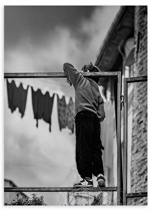 Cuadro fotográfico de un niño de espaldas sobre una estructura metálica, con tonos negro, gris y blanco en una escena urbana nostálgica. Disponible en kudeko.com